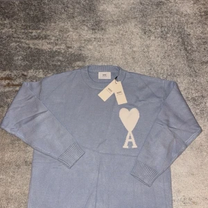 Ljusblå stickad tröja från Ami - Snygg ljusblå stickad tröja från Ami med ett vitt hjärta och bokstaven A på bröstet. Tröjan har rund hals, ribbade muddar och långa ärmar. Perfekt för dig som gillar stilrena detaljer och mjuka material.
