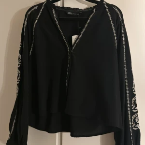 Svart blus med broderade detaljer från Zara - Svart blus från Zara med vita broderade detaljer längs ärmar och axlar. Blusen har en lös passform, långärmade ärmar och ett omlottliknande fall framtill. Materialet är lätt och flowy, perfekt för att ge en chill och snygg vibe.