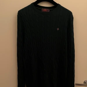 Mörkgrön kabelstickad tröja Morris - Snygg mörkgrön kabelstickad tröja från Morris i 100% merinoull. Tröjan har rund hals med en broderad logga på bröstet. Perfekt för den som gillar klassisk stil med en modern touch. Tröjan är då även rätt ny då jag bara testat i butik innan jag köpte, priset låg då på 1900kr.