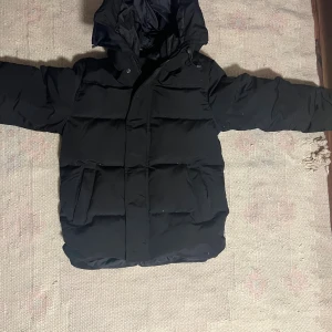 Svart pufferjacka från Canada Goose - Svart pufferjacka från Canada Goose i storlek M. Jackan har huva, stora fickor framtill och dragkedja med knappslå. Materialet är polyester och dun, perfekt för kalla dagar. Snygg och stilren design som håller dig varm hela vintern.