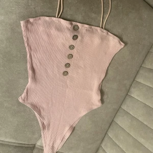 Rosa ribbad body med knappar - Supersnygg ljusrosa body med ribbad struktur och sex dekorativa knappar framtill. Modellen har smala axelband och en stilren, enkel form som ger en modern vibe. Oanvänd 
