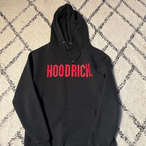 Svart hoodie från Hoodrich med röd text - Svart hoodie från Hoodrich med stor röd logga på bröstet. Klassisk huva med snörning och magficka. Perfekt för en streetwear-look och riktigt skön att bära. Långärmad och i mjukt material.
