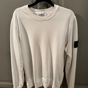 Vit tröja från Stone Island - Vit sweatshirt från Stone Island med klassisk rund hals och ikonisk svart logotyp-patch på vänster arm. Perfekt för dig som gillar stilrena och trendiga plagg. Köpt på NK