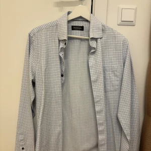 Rutig skjorta Dressmann M - Vit skjorta från Dressmann i premium bomull med rutigt mönster i blått och brunt. Klassisk krage, långärmad och med knappar framtill. Perfekt för dig som gillar stilren och enkel design.