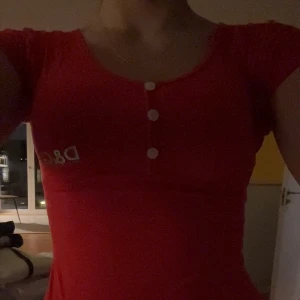 Röd topp från D&G med knappar - Säljer en röd topp från D&G med korta ärmar och v-ringad hals. Toppen har tre vita knappar framtill och en silverfärgad D&G-logga på bröstet. Materialet känns mjukt och stretchigt, perfekt för dig som gillar stilrena detaljer.
