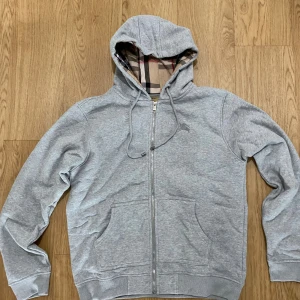 Grå Burberry hoodie  - Snygg grå hoodie från Burberry med klassisk rutig insida i huvan. Tröjan har dragkedja, snörning och broderad logga på bröstet. Tillverkad i mjuk bomull och har ribbade muddar samt två fickor framtill. Perfekt för dig som gillar stilrena detaljer och ikoniskt mönster.