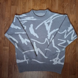 Grå stickad tröja med vitt mönster - Snygg grå stickad tröja med vitt abstrakt mönster. Tröjan har rund hals och ribbade muddar vid ärmslut och nederkant. Den är långärmad och har en loose passform som ger en chill vibe. Perfekt för dig som gillar grafiska detaljer och mjuka material. Skriv gärna om ni vill ha mått😁