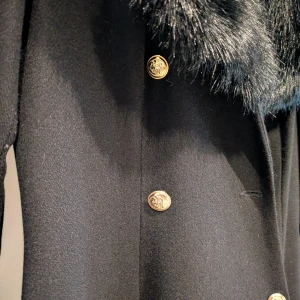Ida Sjöstedt kappa fuskpäls & guldknappar - Snygg svart kappa med bred krage i svart fuskpäls och dekorativa guldknappar framtill. Långärmad och med smickrande passform, perfekt för att ge en lyxig vibe till din outfit. Figursydd. Liten i strl, bra att få plats med ex en stickad tröja under. Dyr i inköp, som ny. Säljer billigt för en Ida Sjöstedt 