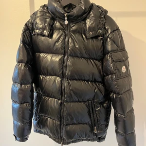 Svart Moncler pufferjacka med huva - Svart pufferjacka från Moncler med glansig finish, huva och dragkedja framtill. Jackan har en ficka med Moncler-logga på ärmen och quiltad design. Perfekt för kalla dagar och har en modern, oversized look. Väldigt osäker på äktheten därav prissättningen!