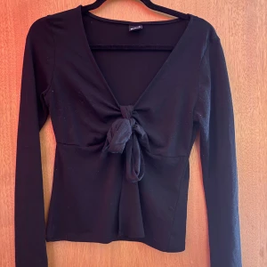 Svart långärmad topp med knyt Gina Tricot - Svart långärmad topp från Gina Tricot med snygg v-ringning och knyt framtill. Modellen är figurnära och har en stilren look som passar till jeans eller kjol. Mjukt material som känns skönt mot huden. Perfekt för dig som gillar enkla men trendiga plagg. nopprig!!
