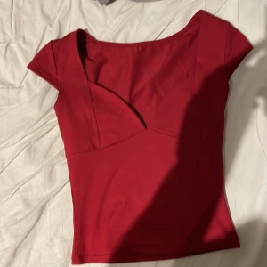 Röd v-ringad topp med kort ärm - Säljer en snygg röd topp med v-ringad hals och korta ärmar. Toppen har en figurnära passform och är tillverkad i ett mjukt, stretchigt syntetmaterial. Perfekt för dig som gillar stilrena och enkla plagg med lite edge.