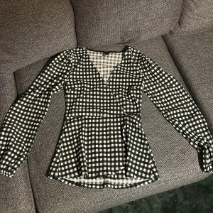 Svartvit rutig omlottblus - Säljer en svartvit rutig omlottblus med långärmade puffärmar och v-ringad hals. Blusen har en figurnära passform och är tillverkad i mjukt, lätt material som känns skönt mot huden. Perfekt för dig som gillar trendiga mönster och snygg siluett.