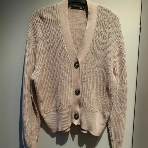 Beige stickad kofta med knappar - Säljer en beige stickad kofta från lager med grovstickad struktur och tre stora knappar framtill. Koftan har lång ärm, ribbad kant vid ärmslut och nederkant samt en djup v-ringning. 