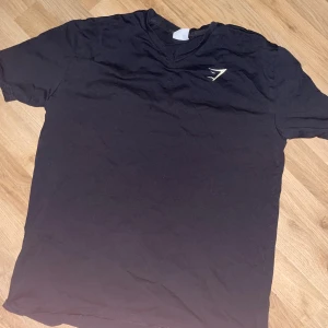 Svart Gymshark t-shirt med v-ringning - Svart t-shirt från Gymshark i storlek S med v-ringad hals och diskret logga på bröstet. Klassisk kortärmad modell i mjukt bomullsmaterial, perfekt för dig som gillar stilren och sportig look.