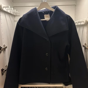 Svart skepparkavaj med bred krage - Stilren mörkblå kappa med tre stora knappar framtill. Jackan har en klassisk rak passform och är långärmad. Enkel design utan mönster eller extra detaljer, vilket gör den lätt att matcha.