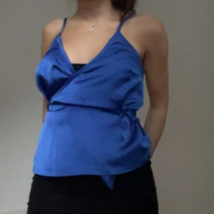 Blå omlott linnetopp i satin - Säljer en snygg blå omlott linnetopp med smala axelband och v-ringning. Toppen har en glansig satinfinish och knyt i sidan för en cool look. Perfekt för dig som gillar att sticka ut med färg och stil. Fråga gärna om fler bilder eller mått!