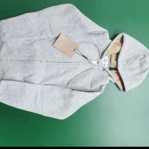 Grå hoodie med dragkedja  - Grå hoodie från Burberry i storlek XS, med klassisk dragkedja framtill och snörning vid huvan. Insidan av huvan har Burberrys ikoniska rutiga tygdetalj. Tröjan är tillverkad i mjuk bomull och har långa ärmar och ribbade muddar för extra komfort. Utan logga vid bröstet
