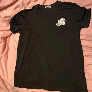 Svart Moncler t-shirt med logga - Svart t-shirt från Moncler med rund hals och korta ärmar. På bröstet sitter två broderade Moncler-loggor i vitt och blått, vilket ger en clean och exklusiv vibe. Materialet är mjuk bomull som känns skönt mot huden. Perfekt för dig som gillar stilrena märkesplagg.