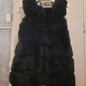 Svart fluffig pälsväst - Säljer en svart pälsväst med riktigt fluffig och mjuk känsla. Västen har en rak och lite längre passform som ger en cool siluett. Perfekt att slänga över en hoodie eller tröja för extra värme och stil. Pälsen är tät och ger ett lyxigt intryck.