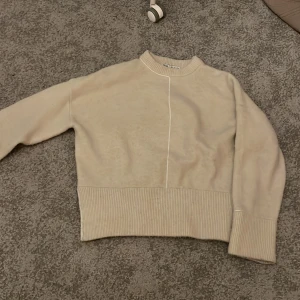 Beige stickad tröja från Zara - Snygg beige stickad tröja från Zara med bred ribbad kant vid midja och ärmslut. Tröjan har en cool asymmetrisk söm framtill och bak, samt rund halsringning. Perfekt för dig som gillar stilrena och enkla plagg med en twist.