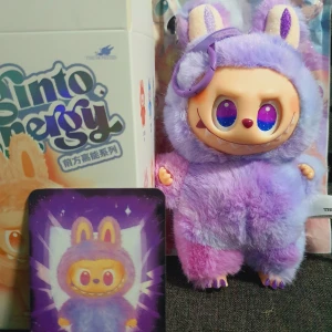 Mjukisdjur Labubu  - Söt och unik samlarfigur från The Monsters, Biginto Energy-serien. Mjuk lila och rosa päls, stora färgglada ögon och kaninöron. Levereras med originalförpackning och samlarkort. Produkten ser ut att vara i nyskick utan synligt slitage. Lycka till 
