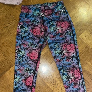 Färgglada träningsbyxor med palmbladsmönster - Säljer ett par leggings med färgstarkt palmbladsmönster i blått, rosa, lila och svart. De har hög midja och snörning i midjan för extra passform. Perfekta för träning eller chill. Materialet är stretchigt och mjukt syntet.