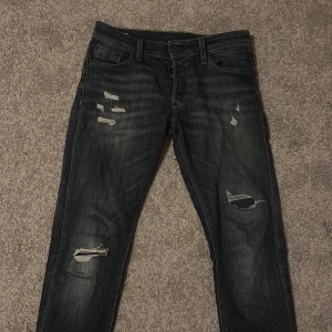 Mörkblå slitna jeans Jack & Jones - Mörkblå jeans från Jack & Jones med slitna detaljer och hål på benen för en cool, avslappnad look. Klassisk femficksmodell med snygga kontrastsömmar och en blå patch bak i midjan. Jeansen har en smal passform och är tillverkade i jeansmaterial.