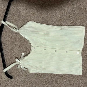 Vit ribbad linnetopp med rosetter - Supersöt vit linnetopp med ribbat mönster och små knappar framtill. Axelbanden är dekorerade med spets och söta rosetter. Perfekt för en somrig look. Strl s