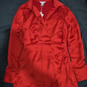 Röd omlottklänning från Amisu - Snygg röd omlottklänning från Amisu i glansigt satinliknande material. Klänningen har lång ärm med manschett och klassisk krage, samt knappar vid ärmslut. Passformen är normal och färgen är intensivt röd. Perfekt för dig som vill sticka ut med en elegant look. Även perfekt för jul eller nyår firandet.