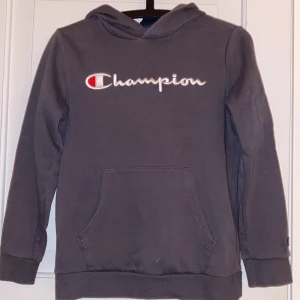Grå hoodie från Champion - Mysig grå hoodie från Champion med broderad logga i vitt och rött på bröstet. Klassisk känguruficka framtill, huva och långa ärmar. Perfekt för chill dagar eller när du vill ha en enkel streetstyle-look.