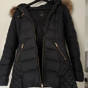 Svart dunjacka Rockandblue med päls - Svart dunjacka från Rockandblue med snygg päls på huvan och guldfärgade dragkedjor. Jackan har quiltade detaljer på sidorna, två fickor med dragkedja och långa ärmar med dragkedja vid mudden. Perfekt för kalla vinterdagar och har en riktigt schysst passform. Tyvärr är dragkedjan sönder, ibland går den att stänga och ibland går den upp.. så det behövs bytas ut om man ska kunna stänga jackan ordentligt. Annars är jackan felfri förutom små slitage. Inköpspris: 4500 kr 