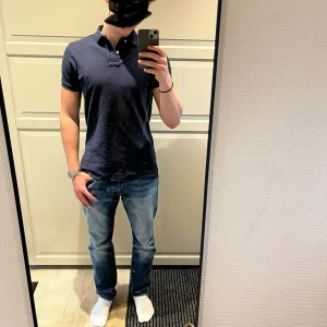 Marinblå pikétröja från Polo Ralph Lauren - Snygg marinblå pikétröja från Polo Ralph Lauren i custom fit. Tröjan har klassisk krage, korta ärmar och två knappar vid halsen. Den ikoniska röda Polo-loggan är broderad på bröstet. Perfekt för dig som gillar stilrena och tidlösa plagg.