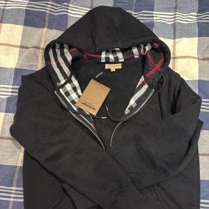 Svart Burberry hoodie med rutig huva - Svart hoodie från Burberry med klassisk rutig insida på huvan i vitt, grått och rött. Tröjan har dragkedja framtill, två fickor och ribbade muddar. Långärmad och med snygga detaljer som logga på bröstet. Perfekt för dig som gillar stilren streetwear.
