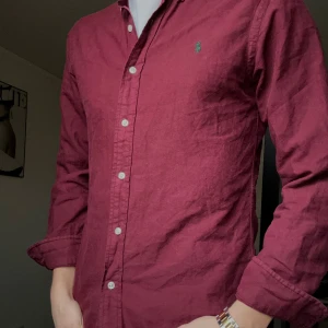 Vinröd skjorta från Ralph Lauren  - Vinröd skjorta från Ralph Lauren i slim fit med klassisk krage och knappar framtill. Skjortan har lång ärm och det ikoniska Polo-logot broderat på bröstet. Materialet är mjuk bomull som känns skönt mot huden. Perfekt för dig som gillar stilrena och snygga plagg.