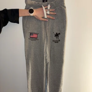 Grå mjukisbyxor från Vinson Polo Club - Snygga grå mjukisbyxor från Vinson Polo Club med broderad logga och amerikansk flagga framtill. Byxorna har elastisk midja, sidofickor och en bakficka. Materialet är mjukt med 50% bomull och 50% viskos, perfekt för chill dagar.