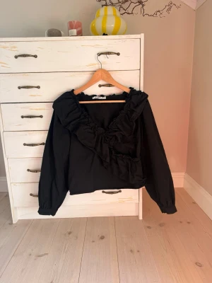 Svart volangblus med v-ringning - Trendig svart blus med djup v-ringning och stora volangdetaljer längs halsringningen. Blusen har långa ärmar med resår vid ärmslut och en lätt puffig siluett. Perfekt för dig som gillar en statement-look. Materialet känns som bomull eller en bomullsblandning. Jätte fin blus som tyvärr blivit för liten för mig 💕