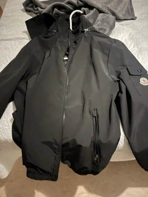 Svart vindjacka med zipper luva - Snygg svart vindjacka  med huva och dragkedja framtill. Jackan har en ficka med lock och M-logga på armen samt en snedställd ficka med dragkedja på framsidan. Tillverkad i ett lätt och vindtätt material, perfekt för blåsiga dagar.