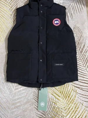 Svart dunväst från Canada Goose  - Snygg svart dunväst från Canada Goose med klassisk logga på bröstet och två stora fickor framtill. Västen har hög krage, tryckknappar och dragkedja. Perfekt för kalla vinterdagar och riktigt stilren look.