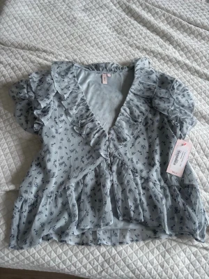 Ljusblå blommig blus med volanger - Supersöt ljusblå blus från Nelly med små mörkblå blommor, volangdetaljer längs v-ringningen och ärmarna samt knappar framtill. Blusen har korta ärmar och är tillverkad i ett lätt, luftigt tyg som känns perfekt för vår och sommar.