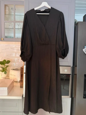 Svart glittrig omlottklänning med puffärm - Stilren svart långklänning med omlott-skärning och v-ringad hals. Klänningen har långa puffärmar med lätt rynk vid axlarna och är tillverkad i ett tyg med subtil glans. Perfekt för dig som gillar en elegant och enkel look.