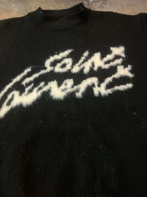 Svart stickad tröja Comme des Garçons - Cool svart stickad tröja från Comme des Garçons med vit fuzzy logga framtill. Tröjan har rund halsringning, långa ärmar och ribbade muddar. Materialet ser ut att vara mjukt och lite luddigt, perfekt för dig som gillar streetwear vibes.