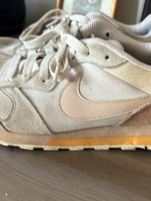 Beige Nike sneakers med mockadetaljer - Snygga sneakers från Nike i beige och ljusbruna toner med klassisk swoosh-logga på sidan. Ovandelen är i mesh och mocka, med vita skosnören och en diskret gul rand på sulan. Perfekta för en avslappnad streetstil.