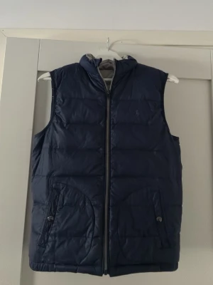 Mörkblå dunväst från Ralph Lauren - Snygg mörkblå dunväst från Ralph Lauren med dragkedja framtill och två fickor med knappstängning. Klassisk quiltad design och diskret logga på bröstet. Perfekt för lager-på-lager och håller dig varm under kyliga dagar.