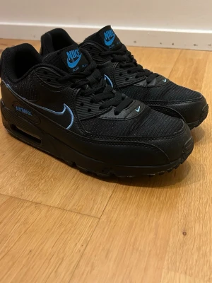 Nike Air Max 90 svart/blå sneakers - Snygga Nike Air Max 90 sneakers i svart med blå detaljer och logga. Skorna har en klassisk låg siluett, synlig Air Max-dämpning i sulan och är tillverkade i en mix av läder och textil. 