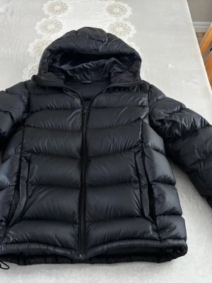 Svart pufferjacka med huva - Snygg svart pufferjacka med huva och dragkedja framtill. Jackan har breda, vadderade paneler och två stora fickor på sidorna. Perfekt för kalla dagar och ger en cool, urban look. Materialet är glansigt och jackan har en avslappnad passform. Bra skick, nyskick. Värmer bra och är bekväm.
