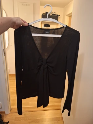 Svart långärmad topp med knytning strl M - Trendig svart långärmad topp från Gina Tricot med djup v-ringning och snygg knytning framtill. Tunn och lätt i materialet, perfekt för att styla med jeans eller kjol. Enkel men ändå iögonfallande design.