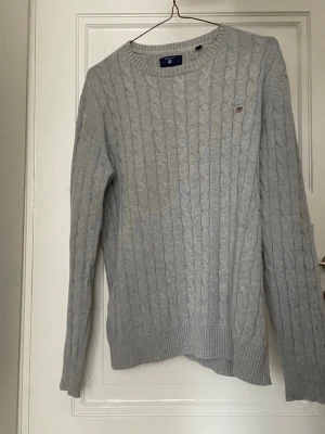 Gant Cableknit - Kabelstickad ljusgrå tröja från Gant med rund halsringning och diskret logga broderad på bröstet. Tröjan har långa ärmar och ribbade muddar vid ärmslut och nederkant. Perfekt för lager-på-lager och klassisk stil.