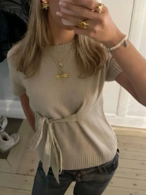 Beige stickad topp med knyt från Zara - Beige stickad topp från Zara med korta ärmar och rund hals. Toppen har en snygg knyt-detalj i midjan som ger en feminin touch. Perfekt att styla med jeans eller kjol för en trendig look.