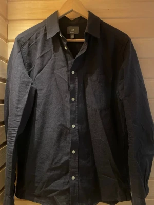 Mörk gråblå skjorta - Klassisk mörk gråblå skjorta från H&M med lång ärm och bröstficka. Skjortan har knappar framtill och vid ärmsluten. Tillverkad i mjuk bomull som känns skön mot huden. Perfekt för dig som gillar en enkel och stilren look.