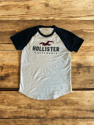  Hollister t-shirt - Snygg vit t-shirt från Hollister med svarta ärmar och kontrastfärgade detaljer. Klassisk raglanmodell med tryckt Hollister-logga och texten 'California' på bröstet. Perfekt till jeans för en avslappnad look.